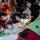 Celebrating the New Year at Task Force Liberty A group of children wearing face masks are seated on the floor engaged in a craft activity, likely making New Year's party hats. In the foreground, a completed green paper party hat with 'New Year!' written on it and decorated with a purple pom-pom and tinsel is prominently visible. The setting appears to be an indoor event or gathering centered around New Year's celebrations.