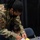Celebrating the New Year at Task Force Liberty A military service member in U.S. Army/Air Force OCP (Operational Camouflage Pattern) uniform, wearing a black face mask, assists a young child with an arts and crafts activity at a table. The service member is leaning over the child, helping with what appears to be a craft project using yellow construction paper and glue. The setting appears to be an indoor event space with dark curtain backdrops.
