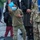 Celebrating the New Year at Task Force Liberty A young boy wearing a blue surgical mask, olive green polo shirt, cargo pants, and bright green/orange Crocs is playing cornhole, mid-throw with a blue bean bag raised in his hand. A military service member in camouflage uniform stands nearby, and several other individuals are visible in the background. The setting appears to be an outdoor gravel area, likely a military base or evacuation/refugee processing site.