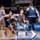 USAFA Men's Basketball Vs Utah State A college basketball game featuring the Air Force Falcons (gray/blue uniforms) playing against an opponent in dark navy uniforms. Players are engaged in an active defensive/offensive play near the perimeter, with Air Force player #3 handling the ball while being guarded. The game is being played in a packed arena with 'Air Force Basketball' branding visible on the court sideline.