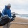 The Harry S. Truman Carrier Strike Group is on a scheduled deployment in the U.S. Sixth Fleet area of operations in support of naval operations to maintain maritime stability and security. A group of volunteers is conducting a beach cleanup on a cloudy, overcast day. The foreground shows a man wearing a blue Adidas hoodie, white cap, and face mask carrying a large black garbage bag filled with collected debris. Several other participants are visible in the background, also wearing gloves and dark clothing, working along the shoreline.