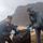 The Harry S. Truman Carrier Strike Group is on a scheduled deployment in the U.S. Sixth Fleet area of operations in support of naval operations to maintain maritime stability and security. Two individuals are participating in a beach cleanup activity on a cloudy, overcast day. They are jointly holding a large black garbage bag, both wearing blue latex gloves for protection. The scene takes place on a sandy beach with a dramatic rocky mountain backdrop and rough sea conditions visible in the background.