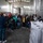 The Harry S. Truman Carrier Strike Group is on a scheduled deployment in the U.S. Sixth Fleet area of operations in support of naval operations to maintain maritime stability and security. A group of approximately 25-30 people wearing face masks are gathered inside what appears to be a winery or brewing facility. A guide or presenter standing with their back to the camera is gesturing toward green-colored equipment, likely explaining the winemaking or brewing process. The industrial space features large stainless steel fermentation tanks and stacked boxed materials.