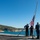The Harry S. Truman Carrier Strike Group is on a scheduled deployment in the U.S. Sixth Fleet area of operations in support of naval operations to maintain maritime stability and security. Three naval personnel are conducting a flag ceremony on what appears to be a ship's deck or pier, raising or lowering a United States flag on a tall flagpole. The scene is set against a coastal backdrop featuring turquoise water, rocky hillsides with vegetation, and clear blue skies. Two individuals are in formal Navy dress uniforms while a third wears a working uniform and is saluting.
