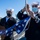 The Harry S. Truman Carrier Strike Group is on a scheduled deployment in the U.S. Sixth Fleet area of operations in support of naval operations to maintain maritime stability and security. Two U.S. Navy sailors in dress blue uniforms and white Dixie cup covers are preparing to raise or lower an American flag using a halyard rope. Both sailors are wearing face masks, suggesting this photo was taken during the COVID-19 pandemic era. The setting appears to be an outdoor naval facility or ship deck with a clear blue sky and hillside visible in the background.