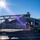 The Harry S. Truman Carrier Strike Group is on a scheduled deployment in the U.S. Sixth Fleet area of operations in support of naval operations to maintain maritime stability and security. A U.S. Navy MH-60S Seahawk helicopter assigned to HSC-11 (Helicopter Sea Combat Squadron 11), tail number 610, is parked on what appears to be an aircraft carrier flight deck. Personnel are performing maintenance or pre-flight checks, with one individual visible on top of the rotor head assembly. The scene is set against a bright sunny sky with mountainous coastline visible in the background.
