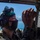 The Harry S. Truman Carrier Strike Group is on a scheduled deployment in the U.S. Sixth Fleet area of operations in support of naval operations to maintain maritime stability and security. A military service member wearing camouflage uniform, hearing protection headset, face mask, and helmet-mounted equipment is performing a technical inspection or maintenance task inside what appears to be a military aircraft or vehicle. The individual is examining or working on a cable or wire connection near a window opening. In the background, a body of water and snow-capped mountains are visible under a clear blue sky.