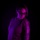 A portrait of a person photographed under dramatic purple/magenta neon lighting against a black background. The subject displays an extensive tattoo sleeve on their right arm featuring floral and face motifs. The lighting creates a moody, artistic aesthetic typical of studio portrait photography.