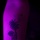 A tattoo on a human arm photographed under ultraviolet (blacklight) illumination, causing the skin to glow purple/magenta. The tattoo depicts a small girl holding a large dandelion flower, with seeds appearing to float away. Small colored marks (red and blue) are also visible near the top of the tattoo, possibly part of the design or reference marks.