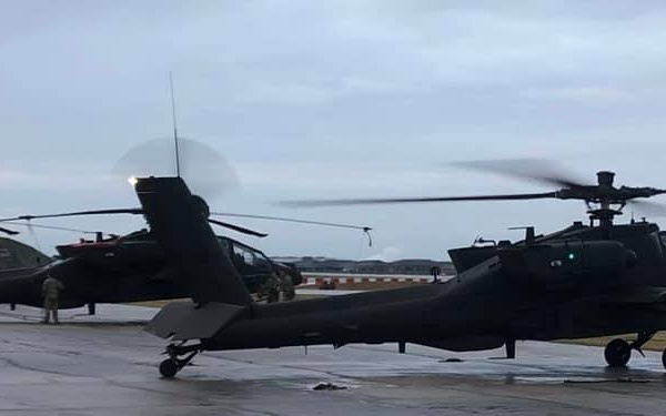 The image shows multiple military attack helicopters, identifiable as AH-64 Apache variants, parked on a wet tarmac under overcast skies. At least one helicopter has its rotors spinning, indicated by motion blur. Several ground crew personnel are visible near the aircraft.
