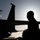 Day by Day with the 492nd Fighter Squadron A dramatic silhouette photograph taken at dusk or dawn showing a person in what appears to be flight gear standing near a military jet aircraft. The backlit composition creates strong contrast between the dark foreground subjects and the bright, hazy sky. The image has an atmospheric, cinematic quality typical of military aviation photography.