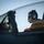 Day by Day with the 492nd Fighter Squadron A military aviator is seated in the cockpit of a jet aircraft, wearing a flight suit, sunglasses, and aviation headset. The canopy is open and shows water droplets on the glass, suggesting recent rain or moisture. A second occupant is partially visible in the front seat of what appears to be a two-seat trainer or combat aircraft.