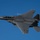 Day by Day with the 492nd Fighter Squadron A U.S. Air Force F-15E Strike Eagle fighter jet is photographed in flight against a clear blue sky. The aircraft is viewed from below at a slight angle, revealing its twin-tail configuration, cockpit, and underwing ordnance. Tail markings indicate it belongs to the 48th Fighter Wing based at RAF Lakenheath, UK, with tail code 'LN' and serial number ending in '605'.