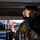Day by Day with the 492nd Fighter Squadron A military or aviation maintenance technician is photographed in close-up while working beneath what appears to be an aircraft. The individual is wearing full personal protective equipment including safety goggles, hearing protection, camouflage uniform, and black gloves. Their uniform shows signs of fluid or grease staining, suggesting active maintenance work.