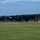 Day by Day with the 492nd Fighter Squadron A military airfield flight line showing multiple fighter jets parked in a row on a taxiway or apron area. Ground crew personnel are visible around the aircraft, suggesting pre-flight or post-flight servicing operations. The scene is set against a backdrop of dense conifer trees with an overcast sky and a windsock visible.