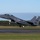 Day by Day with the 492nd Fighter Squadron A U.S. Air Force F-15E Strike Eagle with tail code 'LN' and serial number AF 91-329 is captured during takeoff or landing on a runway. The aircraft is in a two-seat configuration with both crew members visible in the cockpit. The image shows the jet at low altitude with landing gear still deployed and appears to carry external stores/munitions.
