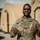 A U.S. Air Force service member named Drummond is posing outdoors in OCP (Operational Camouflage Pattern) uniform, giving a thumbs-up gesture with both hands and smiling broadly. He is wearing sunglasses and his name tape and branch tape are clearly visible on his uniform. The background shows tan/beige military field tents or shelters, suggesting a deployed or field environment.