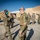 A group of military personnel in OCP (Operational Camouflage Pattern) uniforms are gathered outdoors at what appears to be a forward operating base or deployed location with tan/beige tent structures in the background. Two soldiers in the foreground are performing a fist bump greeting while wearing face masks, suggesting this was taken during the COVID-19 pandemic period. A larger group of uniformed personnel observes in the background.