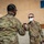Two military personnel in OCP (Operational Camouflage Pattern) uniforms are performing a fist bump greeting, both wearing face masks consistent with COVID-19 era protocols. The setting appears to be a forward operating base or military installation, with tents, a basketball hoop, tires, and a tan concrete wall visible in the background. Additional personnel are partially visible in the background.