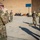 A group of military personnel in OCP (Operational Camouflage Pattern) uniforms are gathered outdoors in what appears to be a forward operating base or military installation. Most individuals are wearing face masks, suggesting this was taken during the COVID-19 pandemic period. A soldier with the name tape 'MILLIGAN' is visible facing another service member in what appears to be a briefing or formation.