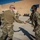 Two U.S. Air Force personnel in OCP (Operational Camouflage Pattern) uniforms are shaking hands at what appears to be a forward operating base or air base in an arid environment. Both individuals are wearing face masks and sunglasses, consistent with COVID-era military operations. Several other uniformed personnel are visible in the background observing the interaction.