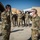 Two military personnel in OCP (Operational Camouflage Pattern) uniforms are exchanging salutes in what appears to be a forward operating base or air base environment. Both individuals are wearing face masks pulled down below their noses, consistent with COVID-19 era protocols. A group of approximately 10-12 similarly uniformed personnel stand at attention in the background near hardened aircraft shelters.