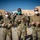 A group of U.S. military personnel in OCP (Operational Camouflage Pattern) uniforms are posed outdoors at what appears to be a forward operating base or air base. Three individuals in the foreground are giving thumbs-up gestures while wearing face masks, with a larger group standing behind them. The center figure's uniform patch reads 'U.S. AIR FORCE' and the name tape reads 'DRUMMOND', and he is holding a challenge coin.