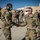 Two military personnel in OCP (Operational Camouflage Pattern) uniforms are shaking hands at what appears to be a forward operating base or military installation. Both individuals and background personnel are wearing face masks, suggesting this occurred during the COVID-19 pandemic period. A group of uniformed service members stands in formation in the background near tan military structures and hangars.
