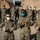 A group of approximately 15-20 U.S. Air Force military personnel posed for a group photo in front of a tan military tent structure. Three individuals in the foreground are giving thumbs-up gestures, with one visibly wearing a name tape reading 'DRUMMOND' and a U.S. Air Force branch tape. All personnel are wearing face masks and OCP (Operational Camouflage Pattern) uniforms, suggesting this was taken during the COVID-19 pandemic era.