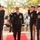 A U.S. Navy officer walks along a red carpet while being rendered a formal side-boy salute by enlisted personnel and fellow officers. The central figure appears to be a Commander or senior officer, identifiable by gold sleeve stripes and multiple service medals, suggesting a formal ceremony such as a change of command or official arrival. The setting appears to be an outdoor naval installation with bollards flanking the red carpet walkway.