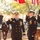 A formal U.S. Navy military ceremony is taking place outdoors, with multiple uniformed naval personnel rendering salutes. The central figure appears to be a senior officer, likely a Commander or Captain, wearing a dress uniform with gold sleeve stripes, medals, and a white combination cover. Several enlisted and officer personnel are saluting in what appears to be a sideboy or honors ceremony.