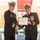 Two U.S. Navy officers in dress uniforms are shaking hands while jointly holding a certificate during a formal military awards ceremony. The certificate is visibly titled 'Meritorious Service Medal' issued by The United States of America. The ceremony appears to be held outdoors at a military installation.
