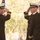 Three U.S. Navy officers in dress uniforms are engaged in a formal military saluting ceremony outdoors. Two officers face each other exchanging salutes while a third stands nearby, with an American flag visible in the background. The setting appears to be a change of command or similar official military ceremony.