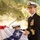 A U.S. Navy officer in dress blues is speaking at a podium during what appears to be a formal military ceremony. He is wearing a white combination cover with gold insignia, multiple service ribbons and medals, and gold sleeve stripes indicating senior officer rank. A blue flag with a white star is visible in the foreground, consistent with a flag officer's pennant or command flag.