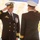 Two U.S. Navy officers in dress blue uniforms are exchanging salutes, likely during a formal military ceremony such as a change of command. Both wear white-topped officer caps and display gold sleeve stripes indicating senior rank. The officer facing the camera wears multiple service ribbons and a large medal on his chest.
