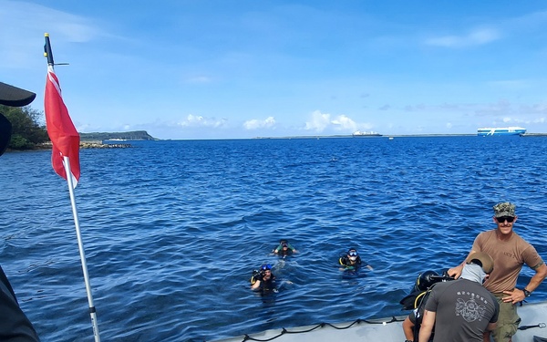 A military diving operation involving personnel from the Republic of Korea and what appears to be U.S. forces at a coastal location. A gray inflatable boat (RHIB) is docked near a pier with diving equipment and personnel aboard, while several divers are visible in the water. A red flag is mounted on the boat, and a Korean military member wearing a face mask and 'SCPO JIN' cap stands in the foreground observing.