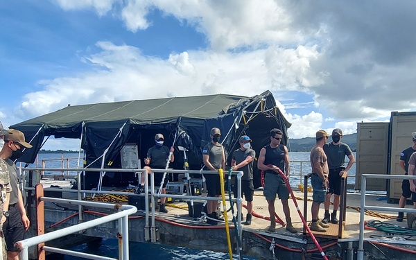 Military personnel from what appears to be a joint U.S. and South Korean operation are gathered on a floating dock or barge near a waterfront facility. A green military tent structure is erected on the platform, and various colored hoses and equipment are visible. The setting appears to be a harbor or naval base, likely in a tropical or subtropical location based on the vegetation and cloud formations.