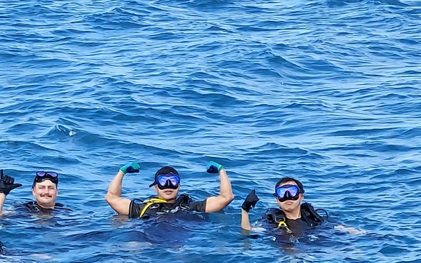Four individuals in scuba diving or snorkeling gear are posing in open blue ocean water, visible from the shoulders up. They are wearing wetsuits, diving masks, and gloves, and appear to be in good spirits, making celebratory gestures. The image was likely taken from a boat or elevated vantage point.