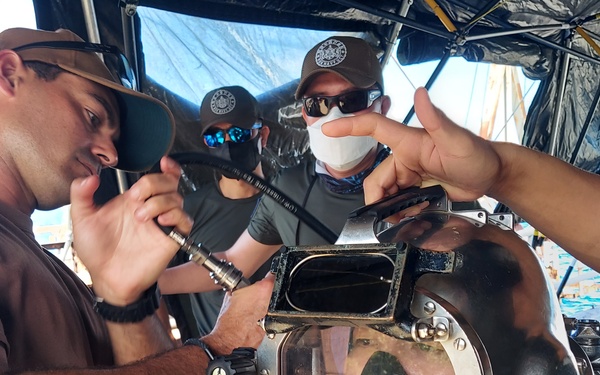 A group of personnel are working on a professional deep-sea diving helmet, likely performing pre-dive checks or maintenance on a vessel or dock. One individual wearing a brown t-shirt with a US flag patch is using a tool on the helmet while others assist. The setting appears to be a maritime environment with ocean visible in the background.