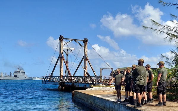 A group of military personnel in matching dark green uniforms and black shorts are gathered on a concrete pier or dock, observing what appears to be a red underwater remotely operated vehicle (ROV) or drone deployed in clear blue-green water. A large metal crane or gantry structure is visible on the dock platform ahead of them, with a naval vessel visible in the background harbor. The setting appears to be a tropical military installation, likely in the Pacific region.
