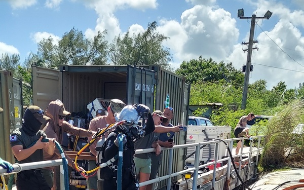 A fully equipped surface-supplied diver in a hard hat helmet and dry suit is being assisted by a support team as they prepare to enter the water from a concrete dive platform. The operation appears to be a professional or military diving operation, with a shipping container serving as a dive support station containing equipment. Multiple umbilical hoses and air lines are visible connecting the diver to the surface support system.