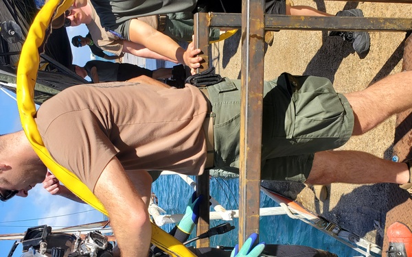 The image shows a diving operation in progress, with personnel assisting a fully equipped scuba diver on what appears to be a metal platform or dive ladder attached to a vessel. Multiple crew members are helping manage equipment including a yellow air hose connected to the diver's gear. The scene takes place over open water under a clear blue sky.