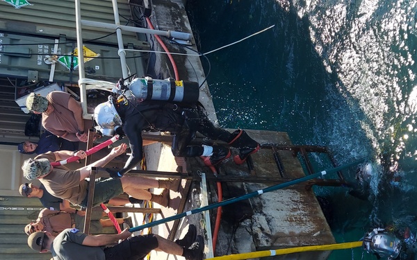 A group of personnel are gathered on a weathered dock or pier structure adjacent to military-style shipping containers, supporting what appears to be a diving operation. At least one diver is visible in the clear turquoise water beside the dock. The scene suggests a military or professional underwater operations exercise or work detail.