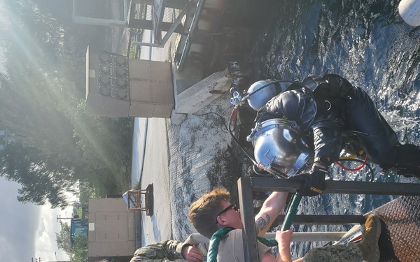 A group of military personnel, appearing to be U.S. Navy sailors in digital camouflage uniforms, are assisting a fully equipped scuba diver near a waterfront. The scene appears to be a dive operation or training exercise, with personnel helping prepare or recover the diver at the water's edge. The image is rotated 90 degrees counterclockwise from its natural orientation.