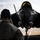 A ground crew member wearing communication headsets observes an F-35 Lightning II stealth fighter jet on a military airfield tarmac. The aircraft is viewed head-on, revealing its distinctive angular stealth geometry, nose landing gear, and open cockpit canopy with a pilot visible inside. Snow-capped mountains are visible in the background, suggesting a high-altitude or northern base location.