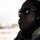 A close-up profile portrait of a man wearing black sunglasses with a small American flag emblem on the frame and black ear protection headphones. He is dressed in what appears to be an olive/sage green flight or tactical jacket. The background is heavily blurred, suggesting an outdoor airfield or tarmac environment.