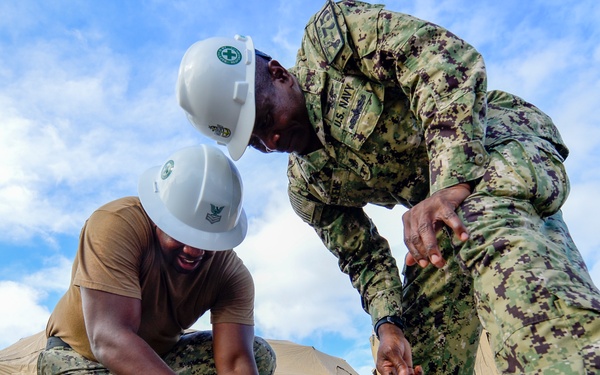 Two U.S. Navy personnel in NWU Type III (AOR-2) camouflage uniforms are working on a ground-level task, with one kneeling and using a pencil or marking tool on a concrete surface. Both individuals are wearing white hard hats with green cross insignia, indicating a medical or construction safety context. Military-style tan/beige tents are visible in the background, suggesting a field hospital or expeditionary base setup.