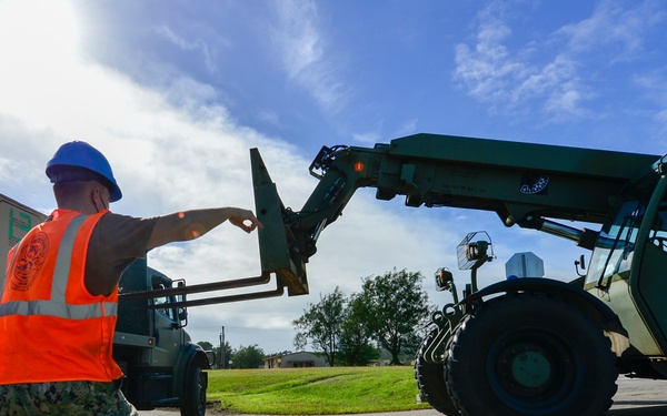 A military service member wearing digital camouflage uniform, orange high-visibility vest, and blue hard hat is directing a large military telehandler/rough terrain forklift on a paved surface. The scene appears to be a logistics or material handling operation, likely at a military installation or disaster relief staging area. At least two other personnel are visible, one operating the forklift and another near a flatbed truck in the background.