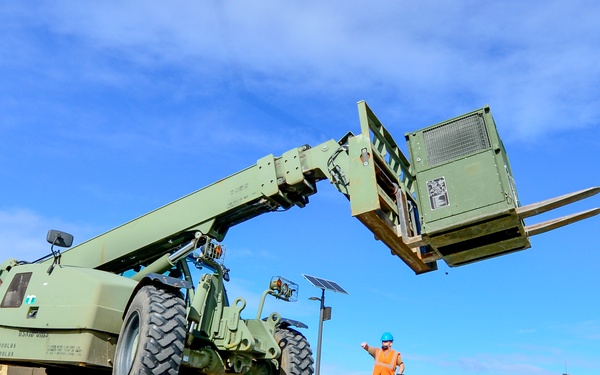 A military telescoping forklift (telehandler) painted in olive drab green is being operated at what appears to be a military installation. The boom arm is extended upward with forks raised, while ground personnel in safety gear direct the operation. Several military personnel and equipment units are visible in the background near a secured perimeter fence.