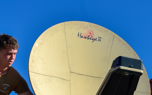 Two military personnel in camouflage uniforms are working on a satellite communications dish labeled 'Hawkeye III' against a clear blue sky. The individuals appear to be installing, adjusting, or maintaining the dish-type antenna system mounted on what appears to be a vehicle or elevated platform. The equipment is consistent with military tactical satellite communication systems.